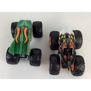 Puede incluir: Dos monster trucks de juguete. Uno es verde con detalles en naranja y un dise&ntilde;o de tent&aacute;culos. El otro es negro con llamas naranjas y amarillas. Ambos tienen neum&aacute;ticos negros grandes.
