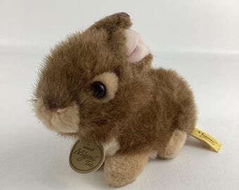 May include: A brown plush rabbit with a white patch on its chest. The rabbit has a pink nose and brown eyes. The rabbit is sitting on a white surface. The rabbit has a tag that says "Mjuni Toys" on it.