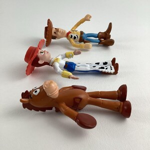 May include: Three Toy Story character figurines: Woody, Jessie, and Bullseye. Woody wears a brown hat, yellow shirt, blue jeans, and brown boots. Jessie has a red hat, white shirt, blue jeans, and cow-print chaps. Bullseye is a brown horse.