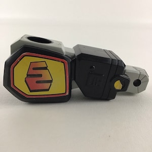 May include: A black and gray plastic toy robot arm with a yellow and red logo that says "S" on the side. The arm is designed to be used with other toys.