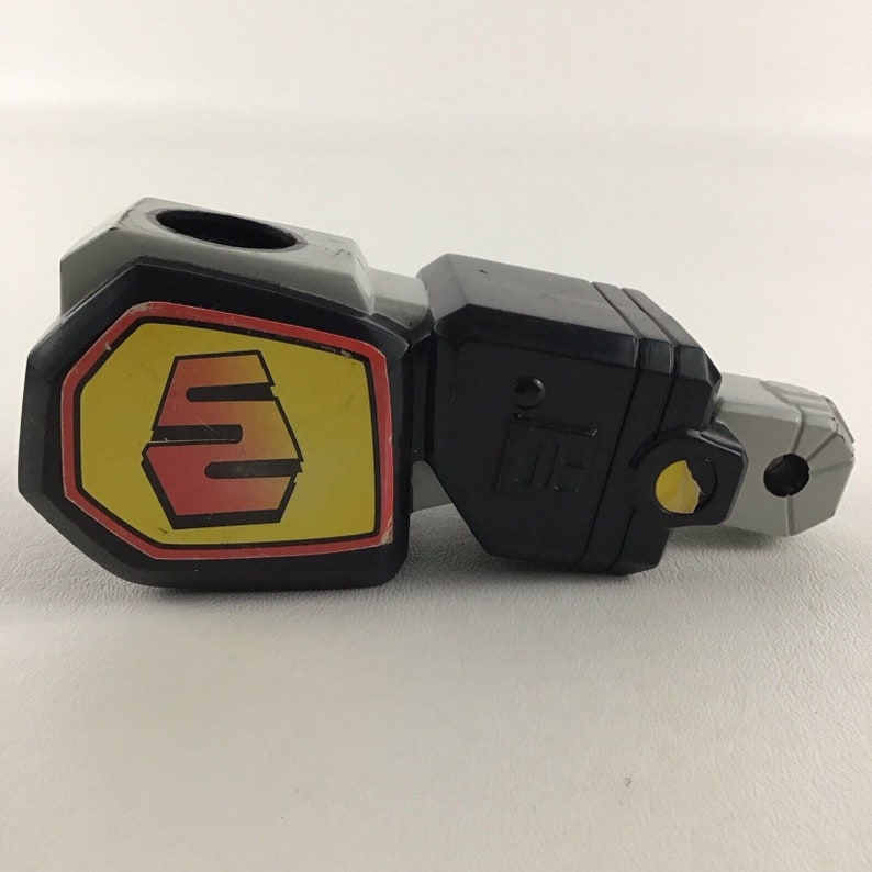 May include: A black and gray plastic toy robot arm with a yellow and red logo that says "S" on the side. The arm is designed to be used with other toys.
