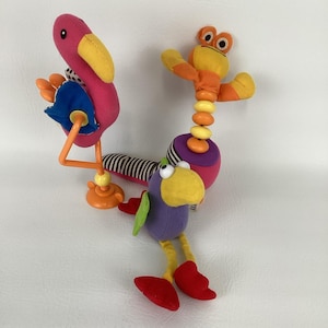 Könnte beinhalten: Ein farbenfrohes Plüsch-Babyspielzeug mit einem rosa Flamingo, einem lila Vogel und einem gelben Frosch. Der Flamingo hat einen gelben Schnabel und orangefarbene Beine. Der lila Vogel hat einen gelben Schnabel und rote Füße. Der Frosch ist gelb und orange.