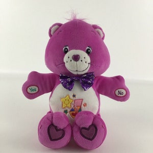 May include: A plush purple Care Bear with a purple bow tie and a white belly with a colorful design. The bear has a yellow star with a smiley face on its belly. The bear has buttons that say "Yes" and "No" on its arms.