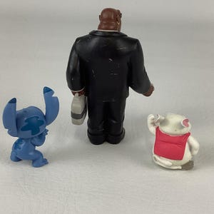 May include: Three small figurines: a blue alien-like character, a man in a black suit, and a white pig wearing a red vest. The figures are on a plain, light-colored surface. The man in the suit is holding a briefcase.