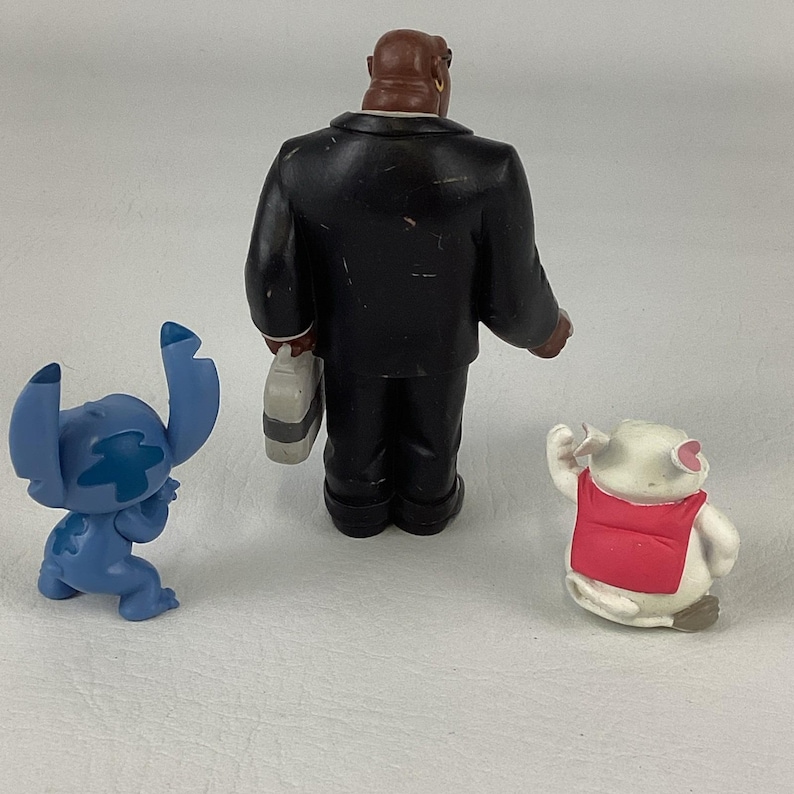 May include: Three small figurines: a blue alien-like character, a man in a black suit, and a white pig wearing a red vest. The figures are on a plain, light-colored surface. The man in the suit is holding a briefcase.