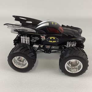May include: A black and silver Hot Wheels monster truck with large, black tires. The truck features the Batman logo in yellow and a red tinted windshield. The vehicle has a unique design with aerodynamic elements.