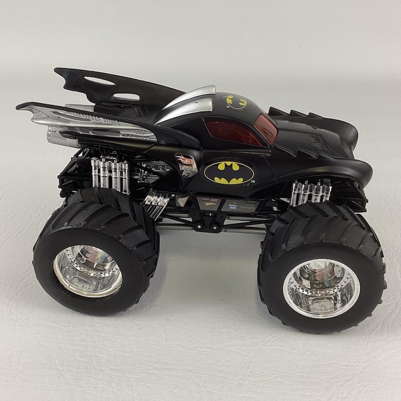 May include: A black and silver Hot Wheels monster truck with large, black tires. The truck features the Batman logo in yellow and a red tinted windshield. The vehicle has a unique design with aerodynamic elements.
