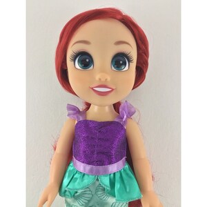 May include: A red-haired doll with blue eyes wearing a purple and green dress. The doll has a sparkly purple top and a green skirt with a seashell pattern.