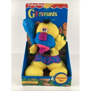 May include: A yellow plush toy with a blue face, pink nose, and a purple tongue. The toy is wearing blue and purple polka dot shorts and is holding a blue balloon. The toy is labelled "Fisher-Price Gr'r'runts" and "Squeeze Me!" and is for ages 2 and up.