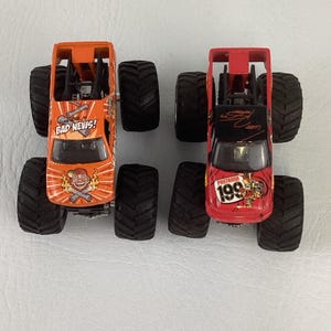 Puede incluir: Dos camiones monstruo de juguete. Uno es naranja con "BAD NEWS!" en el cap&oacute;, el otro es rojo con "PASTRANA 199" en el cap&oacute;. Ambos tienen neum&aacute;ticos negros grandes y chasis detallados.