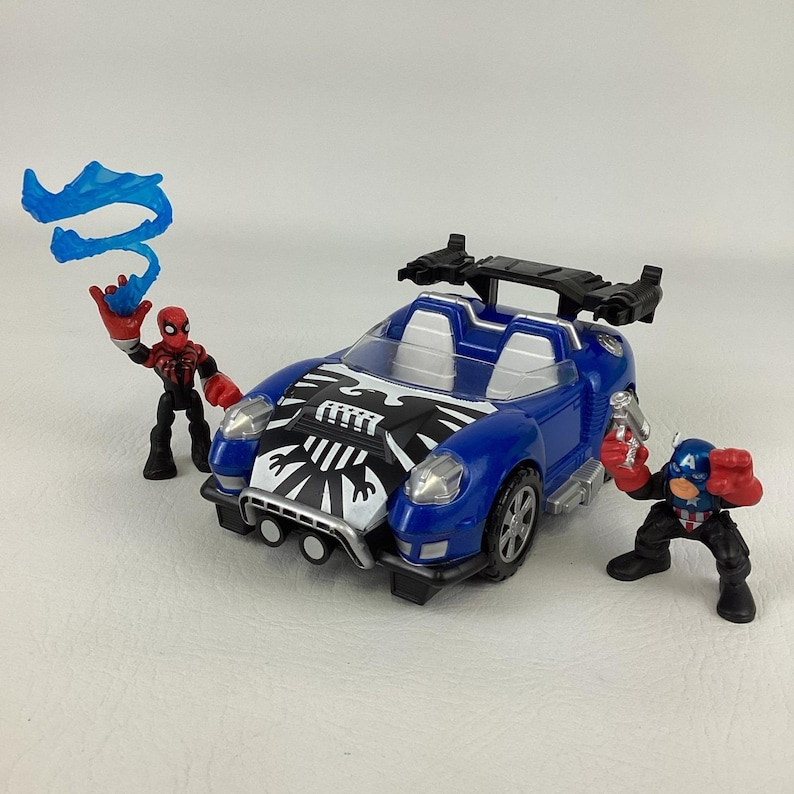 May include: A blue toy car with a black and white design on the hood, flanked by two action figures. One figure is Spider-Man, the other is Captain America. The Spider-Man figure is holding a blue web-like accessory.