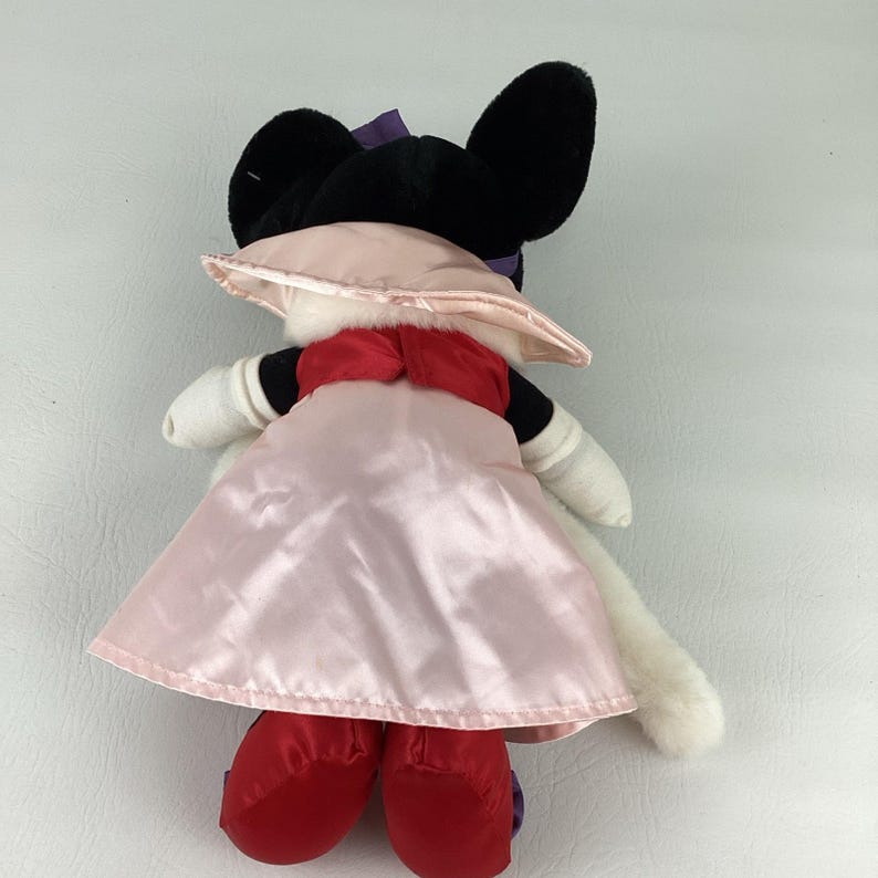 May include: A plush toy featuring a character with black ears, a purple bow, and white arms. The character wears a shiny light pink dress with a wide collar, a red bodice, and red shoes. The back view shows the fabric texture and construction.