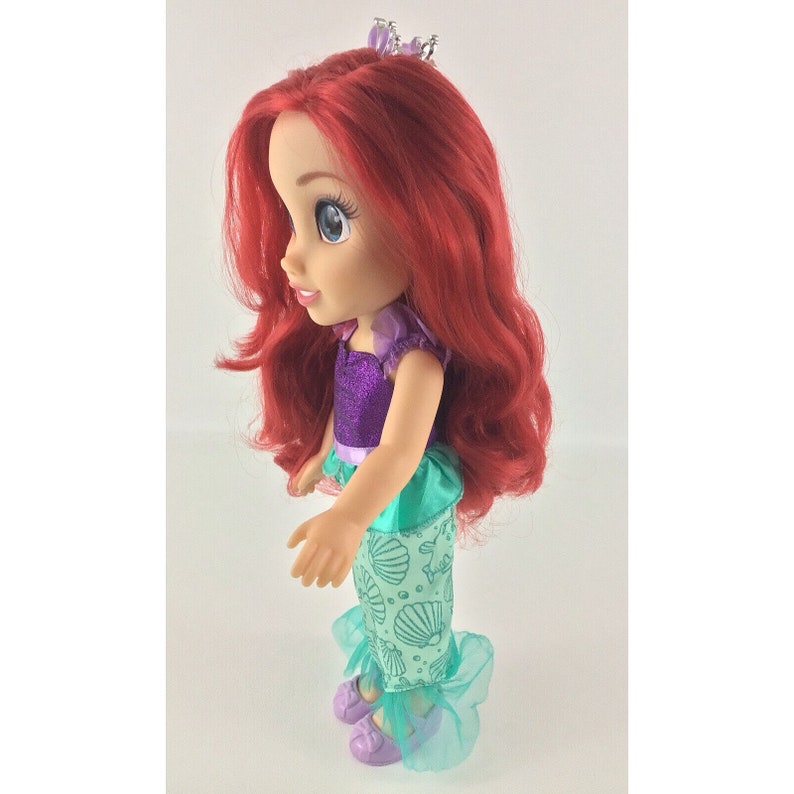 May include: A red-haired doll wearing a purple and green dress with a seashell pattern. The doll is wearing a crown and has a purple and silver glitter top.