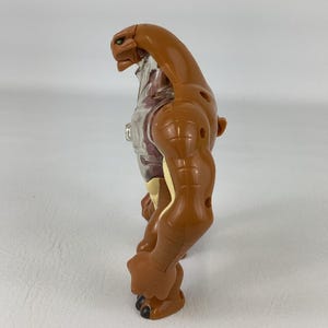 May include: A brown plastic action figure with a smooth, muscular build and a reptilian head. The torso features a translucent clear section revealing internal details, while the limbs are segmented with visible joints and dark brown clawed feet.