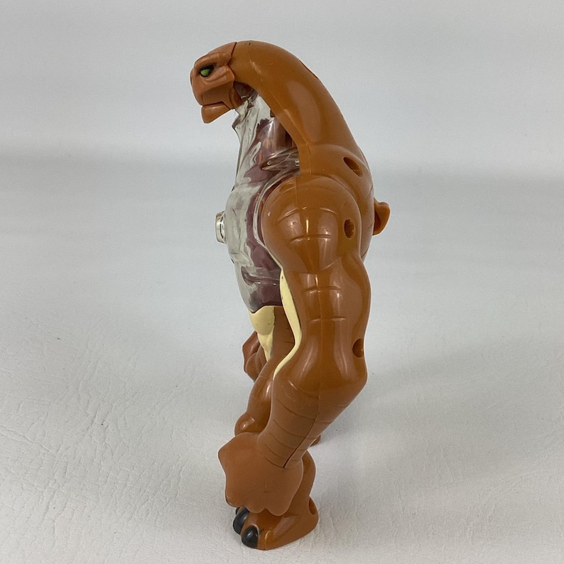 May include: A brown plastic action figure with a smooth, muscular build and a reptilian head. The torso features a translucent clear section revealing internal details, while the limbs are segmented with visible joints and dark brown clawed feet.