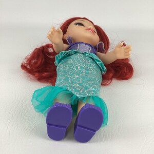 以下が含まれることがあります： 赤い髪の毛をしたアリエルの人形。紫色のトップス、銀色のアクセントが付いたターコイズのスカート、紫色の靴を履いています。人形は腕を広げて横たわっています。