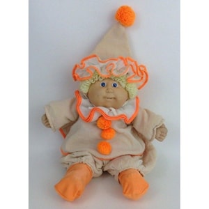 以下が含まれることがあります： キャベツパッチキッズのドールが、ピエロの衣装を着ています。ドールはブロンドの髪と青い目をしており、白とオレンジのピエロの衣装と帽子をかぶっています。