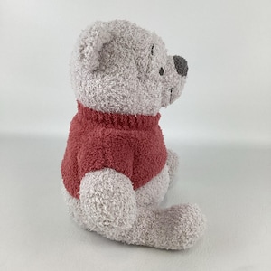 May include: A plush Winnie the Pooh stuffed animal, seated and viewed from the side. The bear is light gray with a dark red sweater. The nose is dark gray, and the eyes are black.