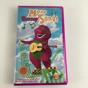 May include: Purple dinosaur named Barney playing a guitar on a background of musical notes. The VHS tape cover reads "More Barney Songs" and "Never Seen On TV".