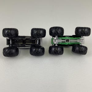 May include: Two toy monster trucks, one silver and one green, are displayed upside down. Both trucks have large black tires and detailed undercarriages. The green truck has a green chassis, while the silver truck has a silver chassis.