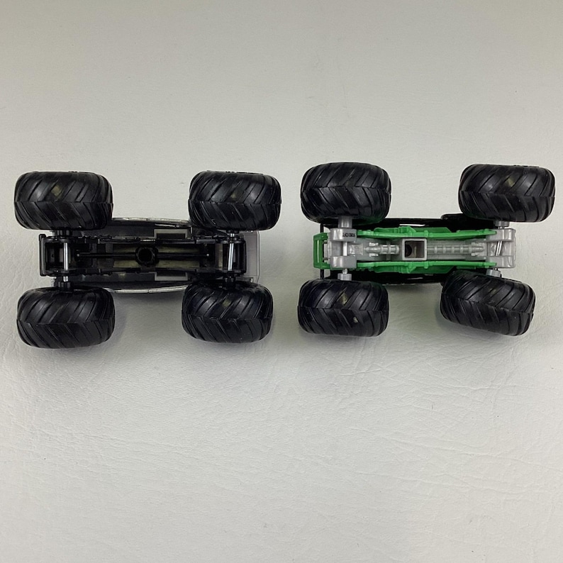 May include: Two toy monster trucks, one silver and one green, are displayed upside down. Both trucks have large black tires and detailed undercarriages. The green truck has a green chassis, while the silver truck has a silver chassis.