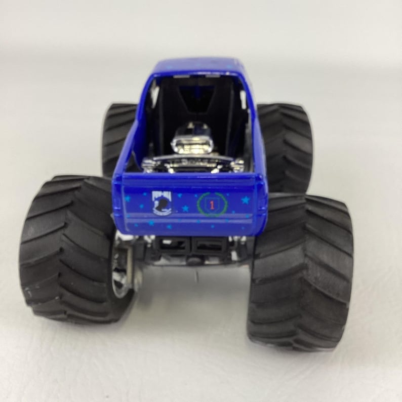 Op de afbeelding: Een blauw monstertruckspeelgoed met grote zwarte banden. De truck heeft een gedetailleerde motor en een rolkooi. De achterkant van de truck heeft een ontwerp met sterren en het nummer 1 in een groene cirkel.