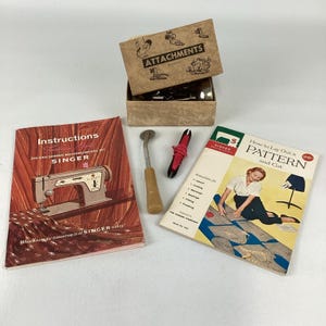 May include: A collection of sewing supplies including a tan cardboard box labeled ATTACHMENTS containing metal parts, a metal tracing wheel with a tan handle, a small black tool with red thread, and two Singer instruction booklets with text.