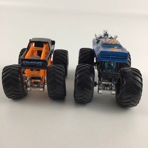 May include: Two monster trucks, one black with orange accents and the word "PREDATOR" on the side, and one blue with orange flames and a silver cage on top.