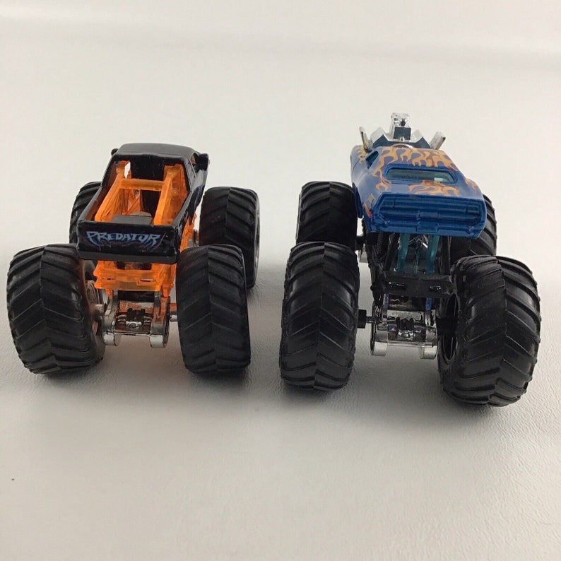 May include: Two monster trucks, one black with orange accents and the word "PREDATOR" on the side, and one blue with orange flames and a silver cage on top.