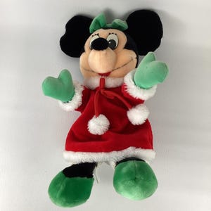 Puede incluir: Un peluche de un personaje de rat&oacute;n de dibujos animados que lleva un vestido de terciopelo rojo con ribete de piel sint&eacute;tica blanca, pompones blancos y un lazo verde. El personaje tiene orejas negras, manoplas verdes y zapatos verdes.