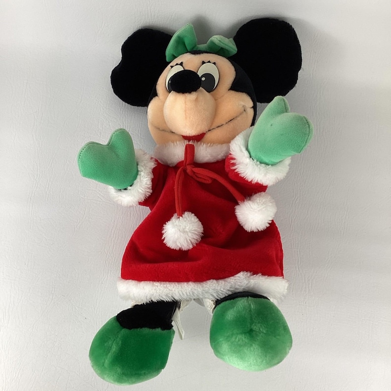 Puede incluir: Un peluche de un personaje de rat&oacute;n de dibujos animados que lleva un vestido de terciopelo rojo con ribete de piel sint&eacute;tica blanca, pompones blancos y un lazo verde. El personaje tiene orejas negras, manoplas verdes y zapatos verdes.