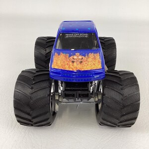 Op de afbeelding: Een blauw monstertruckspeelgoed met grote zwarte banden. De voorkant van de truck heeft een vlammenontwerp met een gespierde figuur. De tekst "TEXAS OFF-ROAD PERFORMANCE" is zichtbaar op de voorruit.