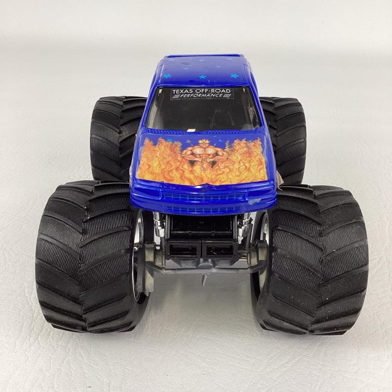 Op de afbeelding: Een blauw monstertruckspeelgoed met grote zwarte banden. De voorkant van de truck heeft een vlammenontwerp met een gespierde figuur. De tekst "TEXAS OFF-ROAD PERFORMANCE" is zichtbaar op de voorruit.