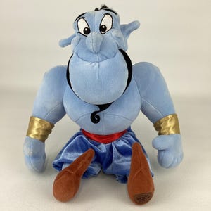 May include: A plush toy of the Genie character with light blue skin, a black beard, and eyebrows. The figure wears gold wrist cuffs, a red waistband, and blue velvet-like pants. The feet are brown, and the character has a large, smiling face.