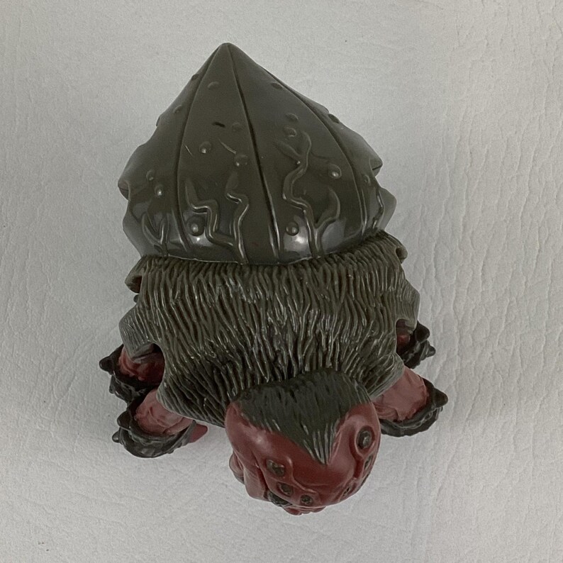 K&ouml;nnte beinhalten: Eine kleine Figur mit einem dunkelbraunen, texturierten, muschelartigen Oberteil mit erhabenen, wellenf&ouml;rmigen Linien. Unter der Schale befindet sich ein dunkelbrauner, fellartiger Mittelteil und eine Basis aus einem rotbraunen Wesen.