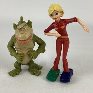 Puede incluir: Una figura de acción de plástico de una criatura verde y escamosa con el pecho color canela y una amplia sonrisa, junto a una figura de acción de plástico de un personaje rubio con un mono rojo, con un zapato verde y otro azul con forma de coche.