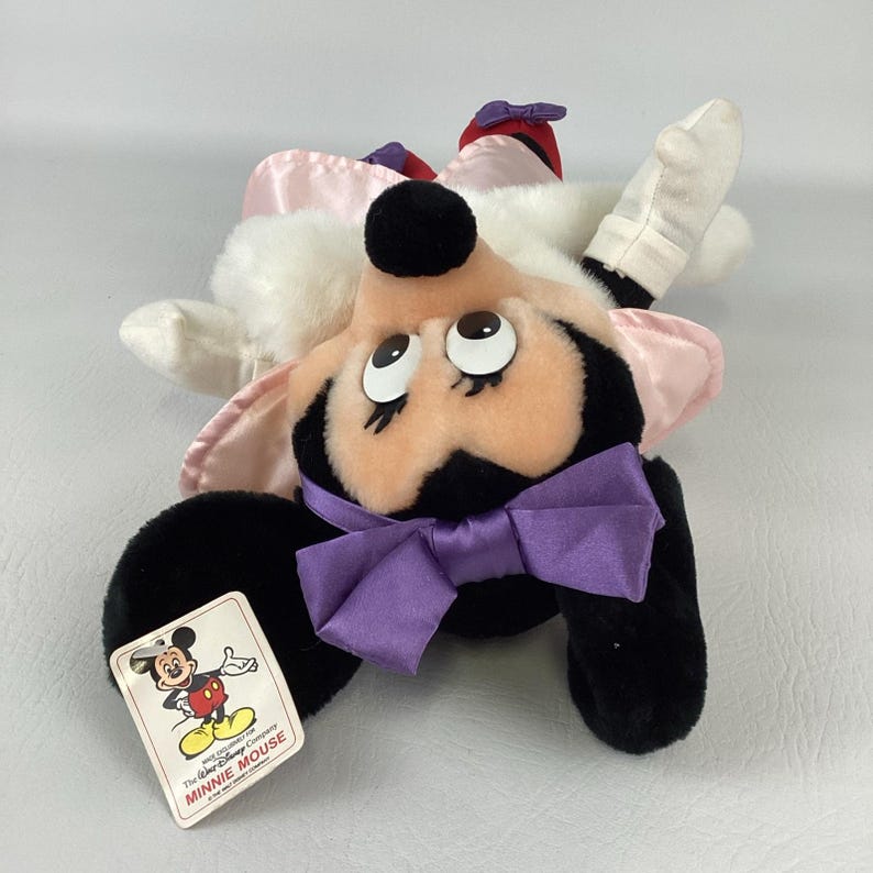 May include: A plush Minnie Mouse toy featuring a black and white body, a peach-coloured face, and a purple satin bow. The character wears a light pink dress and red shoes. A rectangular tag is attached, showing a graphic of Mickey Mouse and text.