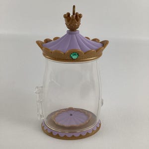Puede incluir: Un tarro de pl&aacute;stico transparente con tapa y base decorativas. La tapa es lavanda con una corona dorada y una gema verde. La base es dorada con un borde festoneado. El tarro tiene un peque&ntilde;o asa lateral.