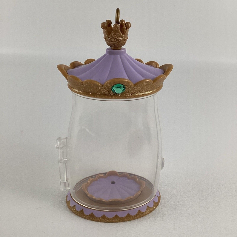 Puede incluir: Un tarro de pl&aacute;stico transparente con tapa y base decorativas. La tapa es lavanda con una corona dorada y una gema verde. La base es dorada con un borde festoneado. El tarro tiene un peque&ntilde;o asa lateral.