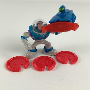 Op de afbeelding: Een Buzz Lightyear actiefiguur in een ruimtepak, met een rode projectiellancer. De figuur is blauw, wit en paars, met een doorzichtige helm. Drie rode, cirkelvormige projectielen met een kroonontwerp liggen ervoor.