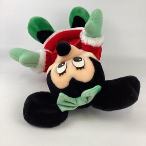 Puede incluir: Un peluche de un personaje con orejas negras, un lazo verde claro y ojos grandes. El personaje viste un conjunto rojo con ribetes de piel sint&eacute;tica blanca en las mangas y detalles verdes en los pies. El juguete est&aacute; colocado boca abajo.