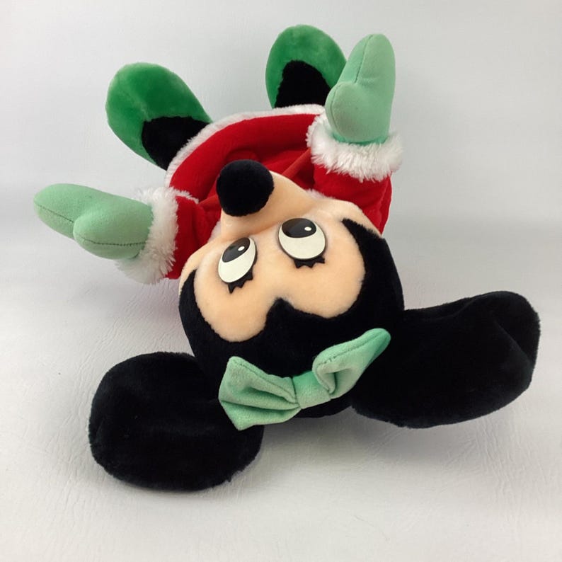 Puede incluir: Un peluche de un personaje con orejas negras, un lazo verde claro y ojos grandes. El personaje viste un conjunto rojo con ribetes de piel sint&eacute;tica blanca en las mangas y detalles verdes en los pies. El juguete est&aacute; colocado boca abajo.