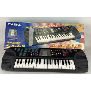 May include: A black Casio SA-65 SongBank keyboard with white keys and a control panel. The keyboard has 37 mini-size keys, 10 Song Bank tunes, 100 tones, and 30 patterns. The box behind the keyboard reads "Musical Information System".