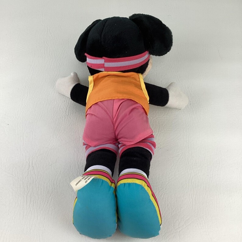 May include: A plush Mickey Mouse toy viewed from the back, featuring black ears, a pink and grey striped headband, an orange vest, pink shorts, and large blue shoes with yellow and red striped accents on the ankles and soles of the feet.