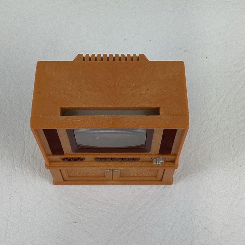 Op de afbeelding: Een kleine, bruine vintage televisie. De tv heeft een rechthoekig scherm, een kast met twee deuren en een decoratieve bovenkant. Het ontwerp omvat knoppen en een luidsprekerrooster. Het is een miniatuurreplica.