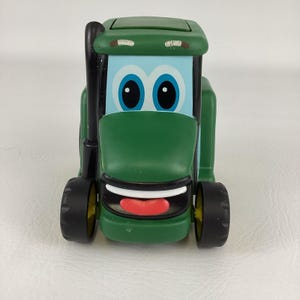 Puede incluir: Un tractor de juguete verde con una cara sonriente, neumáticos negros y un tubo de escape negro. El tractor tiene una cara azul claro con ojos azules grandes y una boca roja. El juguete está diseñado para niños.