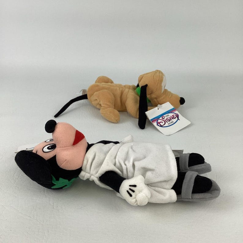 May include: Two plush toys featuring a tan dog with black ears and a green collar, and a black and white character wearing a white robe, green headpiece, and grey sandals. The dog has a white tag attached that reads The Disney Store in blue and red.