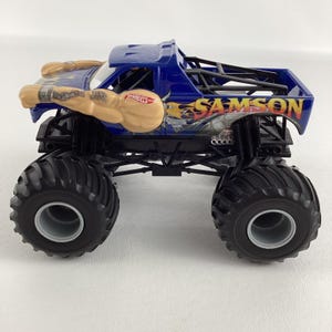 May include: A blue Hot Wheels monster truck toy with the name "SAMSON" in flame-like lettering. The truck features large black tires and a detailed design, including a muscular arm graphic on the side.