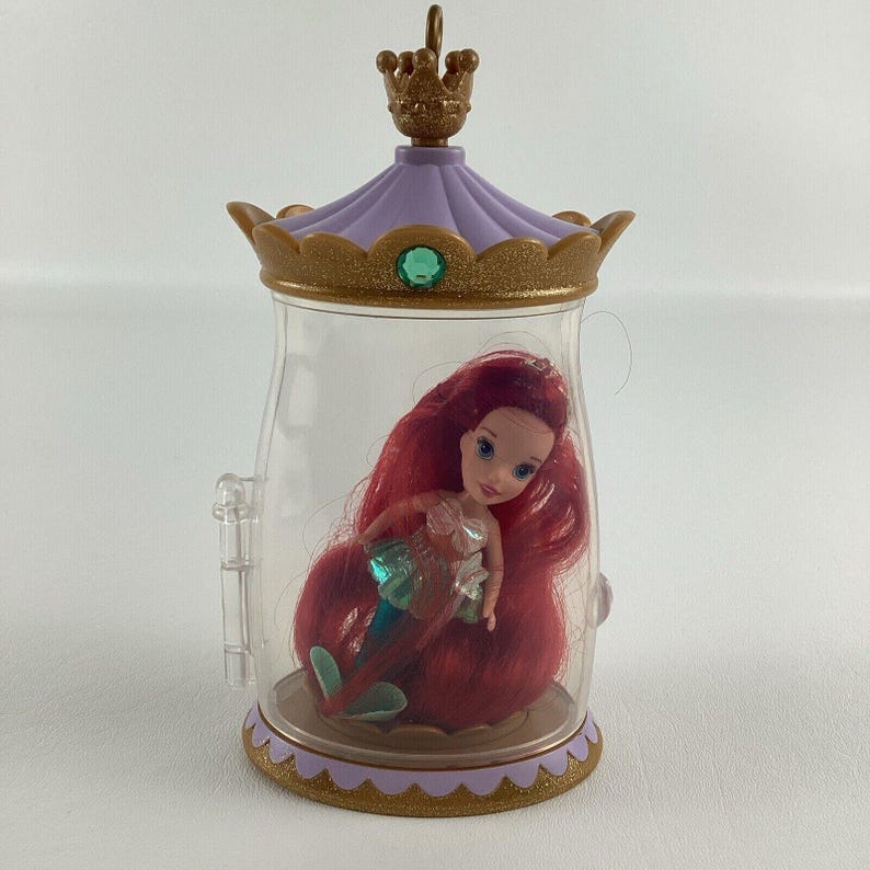 Puede incluir: Mu&ntilde;eca de Ariel dentro de un recipiente de pl&aacute;stico transparente con una tapa dorada y morada en forma de corona. La mu&ntilde;eca tiene el pelo rojo y una cola turquesa. El recipiente tiene una base dorada y un asa transparente.