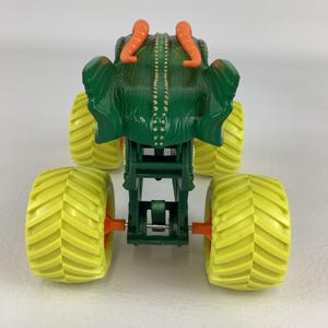 Pu&ograve; includere: Un giocattolo di monster truck verde con un design simile a una creatura sulla parte superiore, con corna arancioni e un corpo strutturato. Il camion ha quattro grandi pneumatici gialli con mozzi arancioni e un telaio verde.